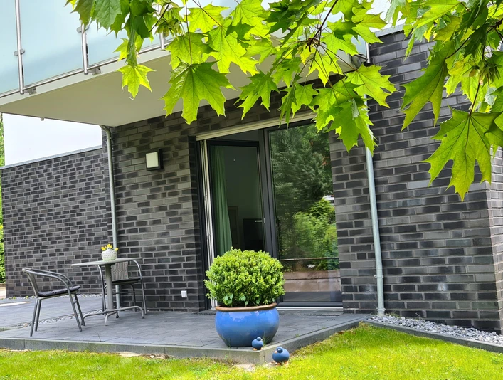 FeWo am Park Außenansicht.jpg Moderne Ferienwohnung mit dunkler Klinkerfassade, Terrasse und Gartenmöbeln in grüner Umgebung.