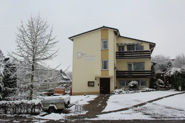 Haus Ostermann 1.JPG Dreistöckiges cremefarbenes Gebäude im winterlichen Umfeld mit Schneebedeckung, Anhänger im Vordergrund.