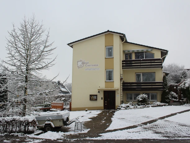 Haus Ostermann 1.JPG Dreistöckiges cremefarbenes Gebäude im winterlichen Umfeld mit Schneebedeckung, Anhänger im Vordergrund.