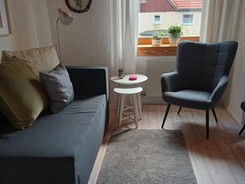 FeWo am Bärenkamp Wohnraum 1.jpg Gemütliches Wohnzimmer mit grauem Sofa, Sessel, Fenster, Vorhängen und Häkchen an der Wand.