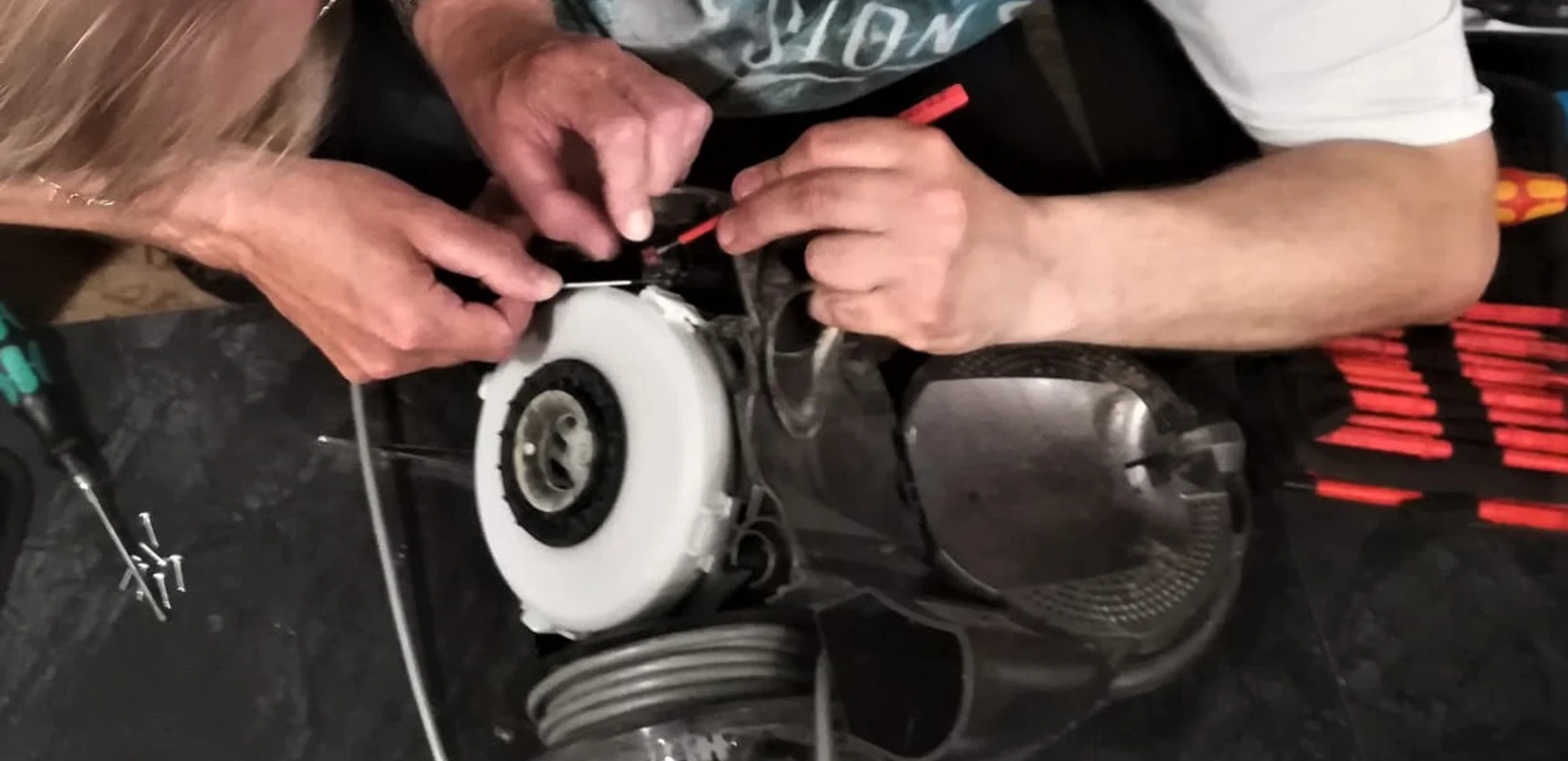 Reparatur Cafe Stemwede Zwei Personen reparieren gemeinsam ein Haushaltsgerät, umgeben von Werkzeug auf einem Tisch.