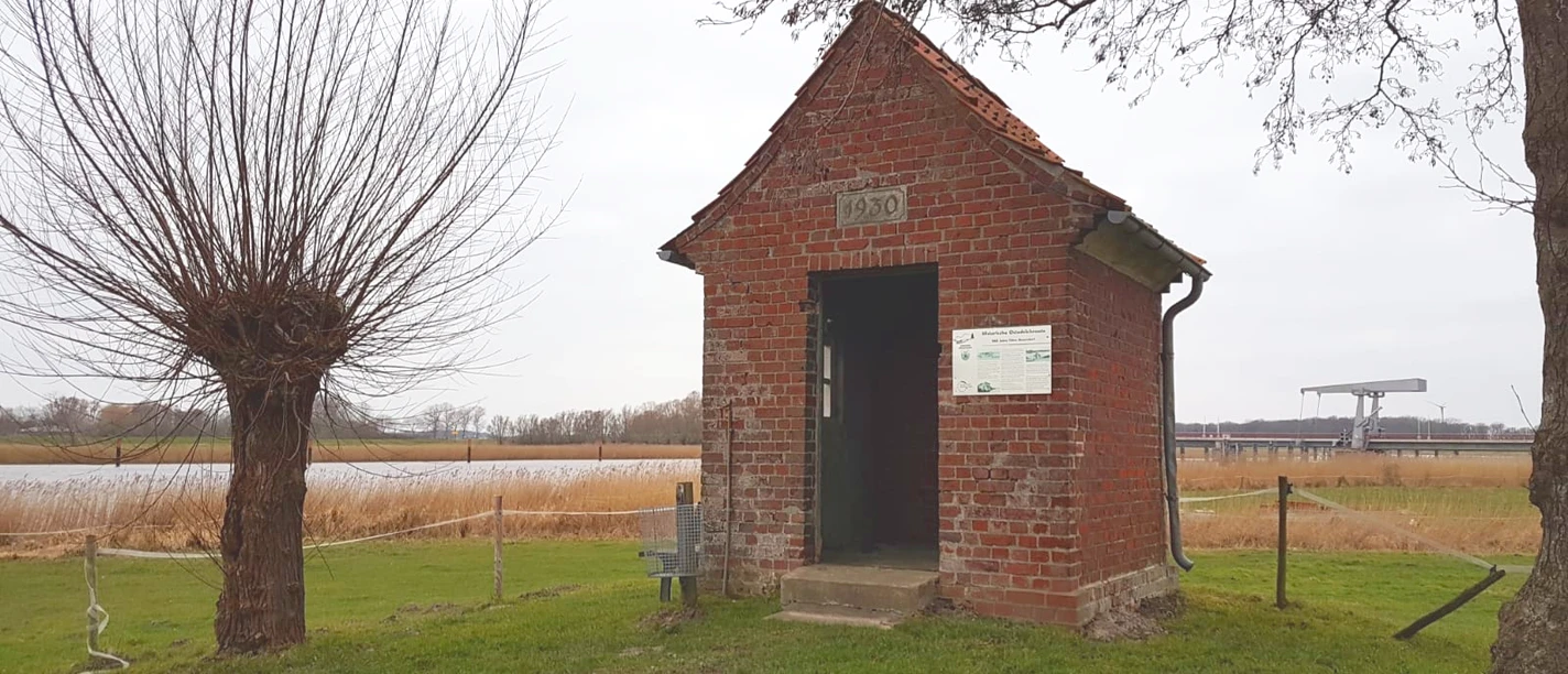 565 Jahre Fähre Geversdorf Kleines Backsteinhäuschen von 1930 am Ostedeich mit Blick auf Flusslandschaft und Brücke in der Ferne.