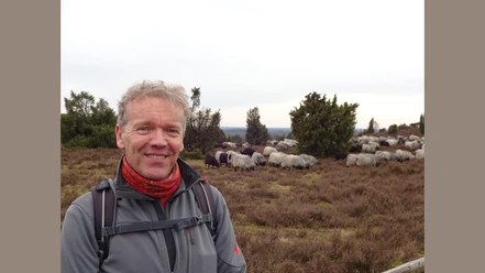 Detlef Grimm_ZNL.JPG Ein lächelnder Mann in Outdoor-Kleidung steht vor einer Herde Schafe in einer bewachsenen Heidelandschaft.