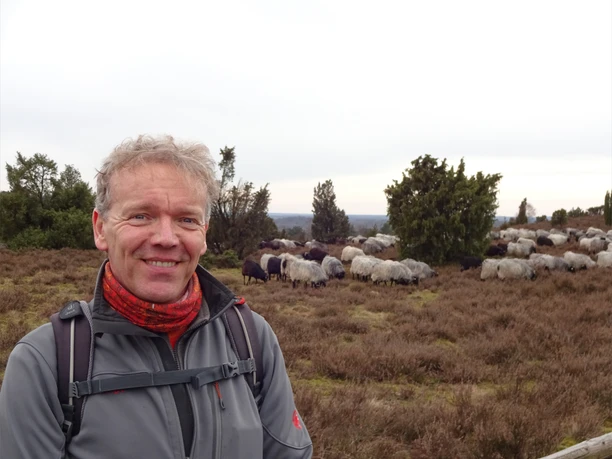 Detlef Grimm_ZNL.JPG Ein lächelnder Mann in Outdoor-Kleidung steht vor einer Herde Schafe in einer bewachsenen Heidelandschaft.