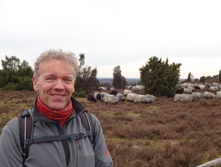Detlef Grimm_ZNL.JPG Ein lächelnder Mann in Outdoor-Kleidung steht vor einer Herde Schafe in einer bewachsenen Heidelandschaft.