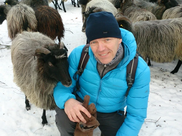 Detlef Grimm_ZNL_1.JPG Ein Mann in leuchtend blauer Jacke kniet im Schnee, umgeben von grauen Schafen, und lächelt sanft in die Kamera.