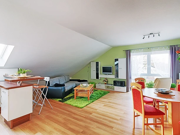 Ein heller Wohnbereich mit grauem Sofa, TV-Ecke, Esstisch, schräger Decke und großen Fensterfläche.