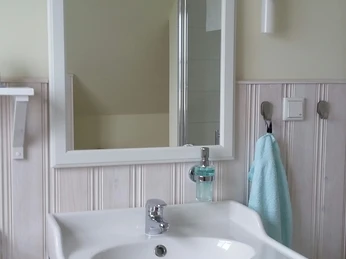 Haus Nordlicht Badezimmer.jpg Modernes Badezimmer mit weißem Waschtisch, Spiegel und hellem Wandpaneel. Handtuch schimmert türkis.