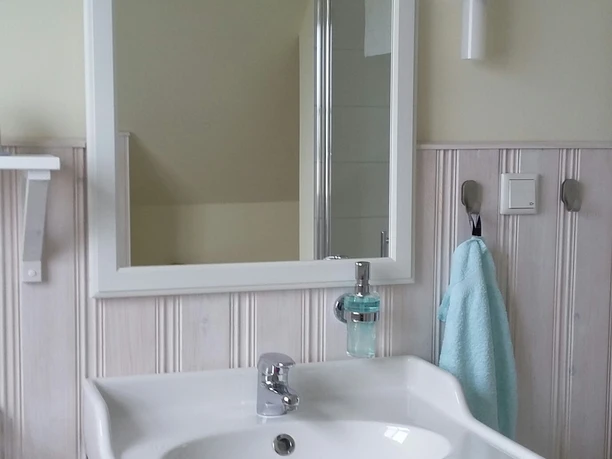 Haus Nordlicht Badezimmer.jpg Modernes Badezimmer mit weißem Waschtisch, Spiegel und hellem Wandpaneel. Handtuch schimmert türkis.