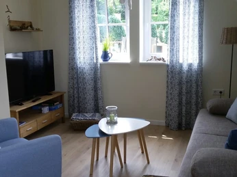 Haus Nordlicht Wohnraum.jpg Wohnzimmer mit hellem Holzfußboden, Sitzgarnitur, Flachbildfernseher und zwei Fenstern mit Vorhängen.