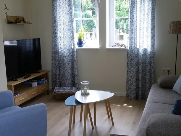 Haus Nordlicht Wohnraum.jpg Wohnzimmer mit hellem Holzfußboden, Sitzgarnitur, Flachbildfernseher und zwei Fenstern mit Vorhängen.