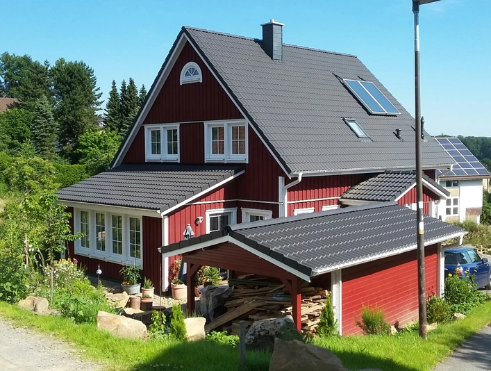 Ein rotes Holzhaus mit grauem Dach steht inmitten grüner Bäume und Sträucher in einer ruhigen Gegend.