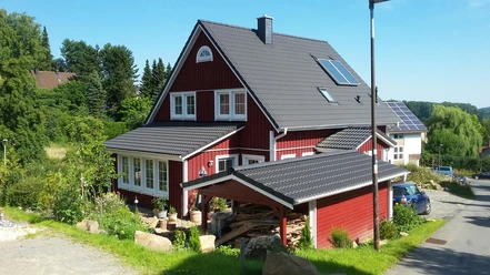 Haus Nordlicht Außenansicht.jpg Ein rotes Holzhaus mit grauem Dach steht inmitten grüner Bäume und Sträucher in einer ruhigen Gegend.