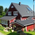Haus Nordlicht Außenansicht.jpg Ein rotes Holzhaus mit grauem Dach steht inmitten grüner Bäume und Sträucher in einer ruhigen Gegend.