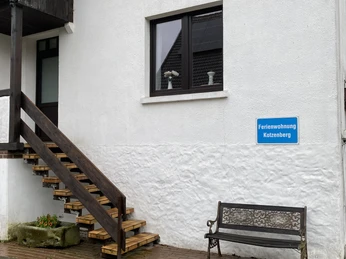 Weiße Ferienwohnung mit Holztreppe, Fenster nach rechts. Bank und Blumenbeet davor auf dem Gehweg.