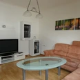 Ferienwohnung "Urlaub in Belle" Wohnbereich 2.jpg Wohnbereich mit beiger Eckcouch, Glastisch, Flatscreen-TV, helle Holzböden und Waldpanorama-Bild an der Wand.