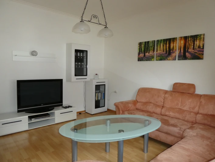 Ferienwohnung "Urlaub in Belle" Wohnbereich 2.jpg Wohnbereich mit beiger Eckcouch, Glastisch, Flatscreen-TV, helle Holzböden und Waldpanorama-Bild an der Wand.
