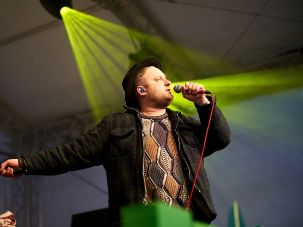 c/o pop in Cologne A singer in a plaid coat and black hat sings into a microphone on a stage with yellow lighting.