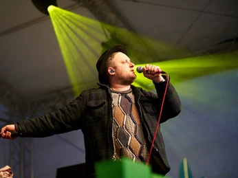 c/o pop in Köln Ein Sänger in einem karierten Mantel und schwarzem Hut singt in ein Mikrofon auf einer Bühne mit gelber Beleuchtung.A singer in a plaid coat and black hat sings into a microphone on a stage with yellow lighting.