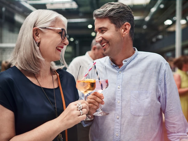 Fine Food Days Cologne A woman and a man look at each other with a laugh on their faces and toast each other with a drink.