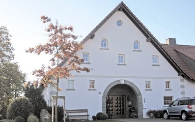ahlsen-reineberg-altes-amtshaus.jpg Historisches Fachwerkhaus mit markantem Giebel, flankiert von einem jungen Baum und geparktem Auto.