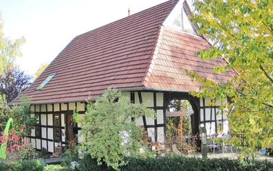 ahlsen-reineberg-heuerlingshaus.jpg Ein Fachwerkhaus mit rotem Ziegeldach umgeben von Herbstlaub und grünem Buschwerk im Vordergrund.