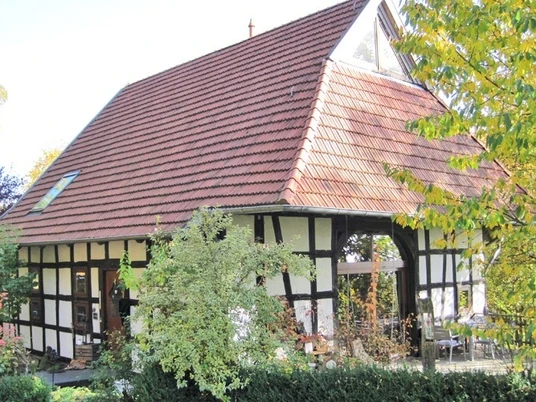 ahlsen-reineberg-heuerlingshaus.jpg Ein Fachwerkhaus mit rotem Ziegeldach umgeben von Herbstlaub und grünem Buschwerk im Vordergrund.