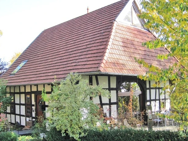 ahlsen-reineberg-heuerlingshaus.jpg Ein Fachwerkhaus mit rotem Ziegeldach umgeben von Herbstlaub und grünem Buschwerk im Vordergrund.