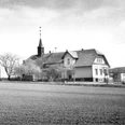 broederhausen-schule-broederhausen-alt.jpg Eine historische Schule mit einem Glockenturm, umgeben von kahlen Bäumen auf einem weiten Feld.