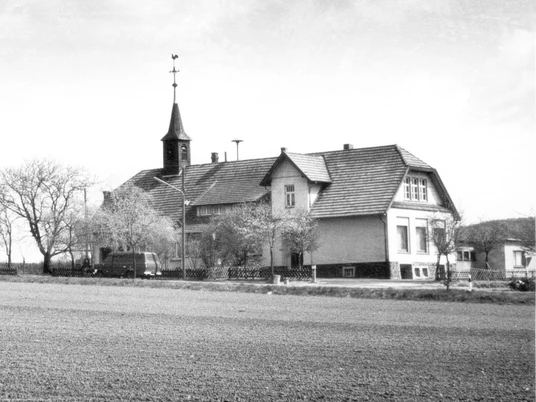 broederhausen-schule-broederhausen-alt.jpg Eine historische Schule mit einem Glockenturm, umgeben von kahlen Bäumen auf einem weiten Feld.