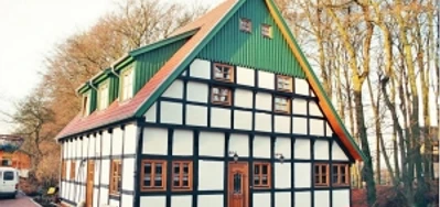 broederhausen-fam-steinmann.jpg Fachwerkhaus mit rotem Satteldach und grüner Giebelwand, umgeben von Bäumen und gepflastertem Hof.