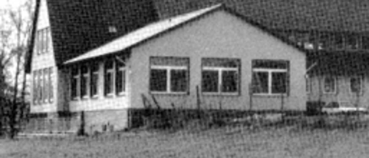 buettendorf-dorfgemeinschaftsraum.jpg Ein schlichtes Gebäude mit Satteldach, großen Fenstern und anliegendem Rasen streckt sich in ländlicher Umgebung.