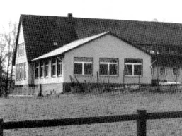 buettendorf-dorfgemeinschaftsraum.jpg Ein schlichtes Gebäude mit Satteldach, großen Fenstern und anliegendem Rasen streckt sich in ländlicher Umgebung.