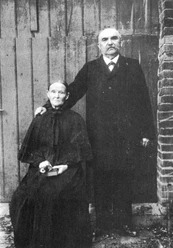 buettendorf-windmuehle-buettendorf.jpg Ein älteres Paar vor einer Holzwand; der Mann stehend, die Frau sitzend mit Blumen in der Hand.