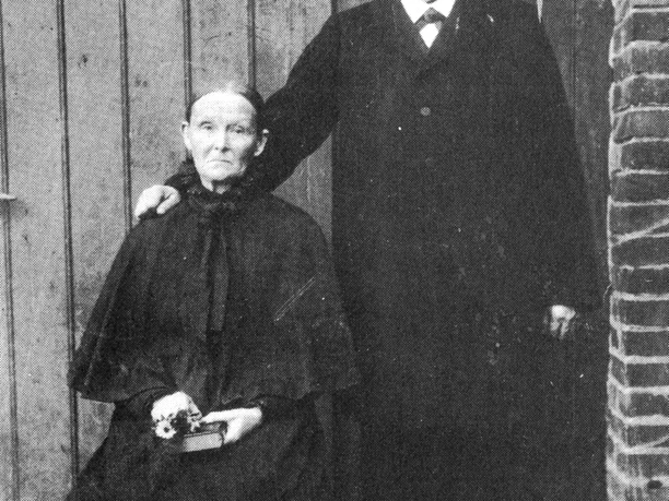 buettendorf-windmuehle-buettendorf.jpg Ein älteres Paar vor einer Holzwand; der Mann stehend, die Frau sitzend mit Blumen in der Hand.