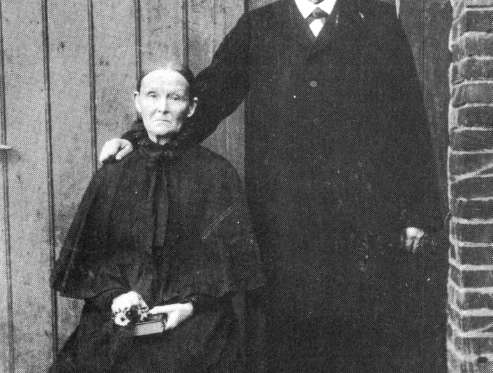 buettendorf-windmuehle-buettendorf.jpg Ein älteres Paar vor einer Holzwand; der Mann stehend, die Frau sitzend mit Blumen in der Hand.