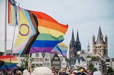 Cologne Pride Rainbow flags fly in the foreground, while Cologne Cathedral can be seen prominently in the background. People are celebrating Cologne Pride.