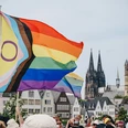 Cologne Pride Rainbow flags fly in the foreground, while Cologne Cathedral can be seen prominently in the background. People are celebrating Cologne Pride.