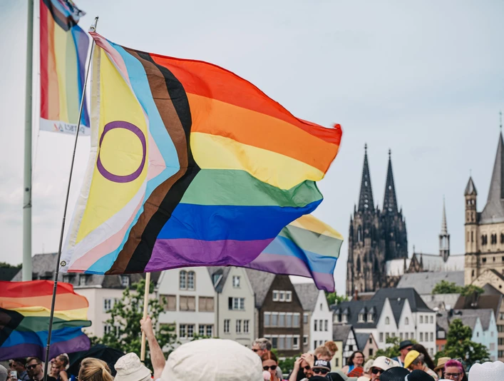 Cologne Pride Regenbogenfahnen wehen im Vordergrund, während der Kölner Dom markant im Hintergrund zu sehen ist. Menschen sind dabei, den Cologne Pride zu feiern.Rainbow flags fly in the foreground, while Cologne Cathedral can be seen prominently in the background. People are celebrating Cologne Pride.
