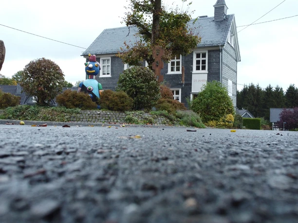 KKH Graues Fachwerkhaus mit Schieferdach, bunte Skulpturen und herbstliches Gebüsch im Vordergrund.