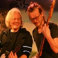 Zwei Musiker auf der Bühne, lächelnd im Scheinwerferlicht, einer spielt Gitarre, der andere Bass.