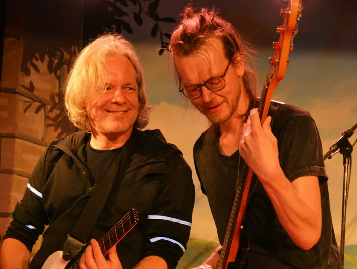 Zwei Musiker auf der Bühne, lächelnd im Scheinwerferlicht, einer spielt Gitarre, der andere Bass.