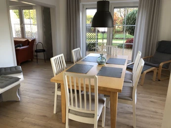 Esszimmer mit Holztisch, weißen Stühlen und großer Fensterfront. Blick auf grünen Garten.