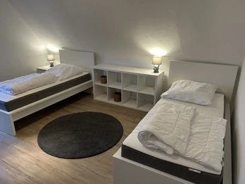 Schlafzimmer_senne-appartementsCC-BY-SA_Teutoburger_Wald_Stadt_Schloss_Holte-Stukenbrock.jpg Zwei Einzelbetten mit weißen Laken, Nachttischlampen und einer Ablage, in einem Zimmer mit Holzfußboden.