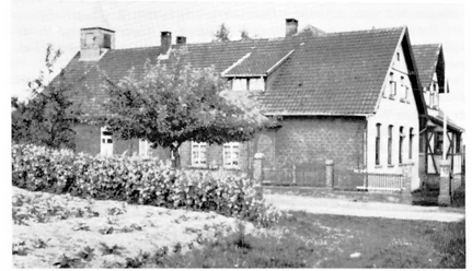 holsen-volksschule-musikschule.jpg Schwarz-Weiß-Foto eines traditionellen Backsteingebäudes mit Satteldach und kleiner grüner Vorplatz.