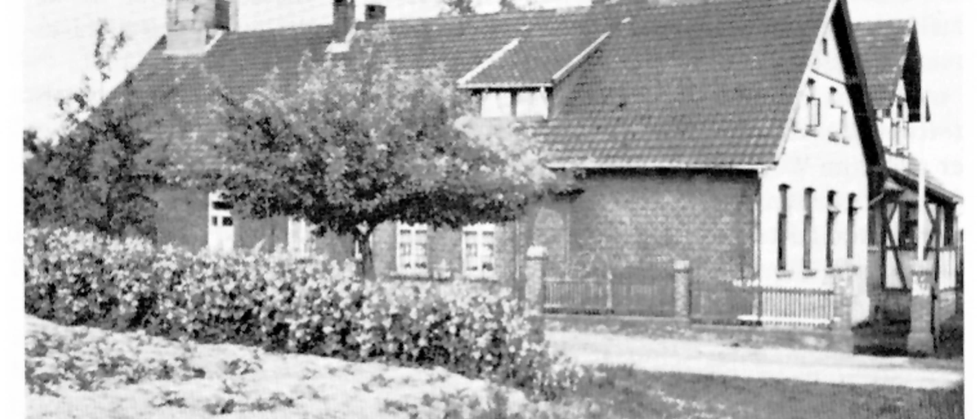 holsen-volksschule-musikschule.jpg Schwarz-Weiß-Foto eines traditionellen Backsteingebäudes mit Satteldach und kleiner grüner Vorplatz.