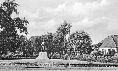 huellhorst-kriegerehrenmal-alt.jpg Kriegerdenkmal in Hüllhorst, umgeben von zwei traditionellen Fachwerkhäusern und Bäumen bei klarem Himmel.