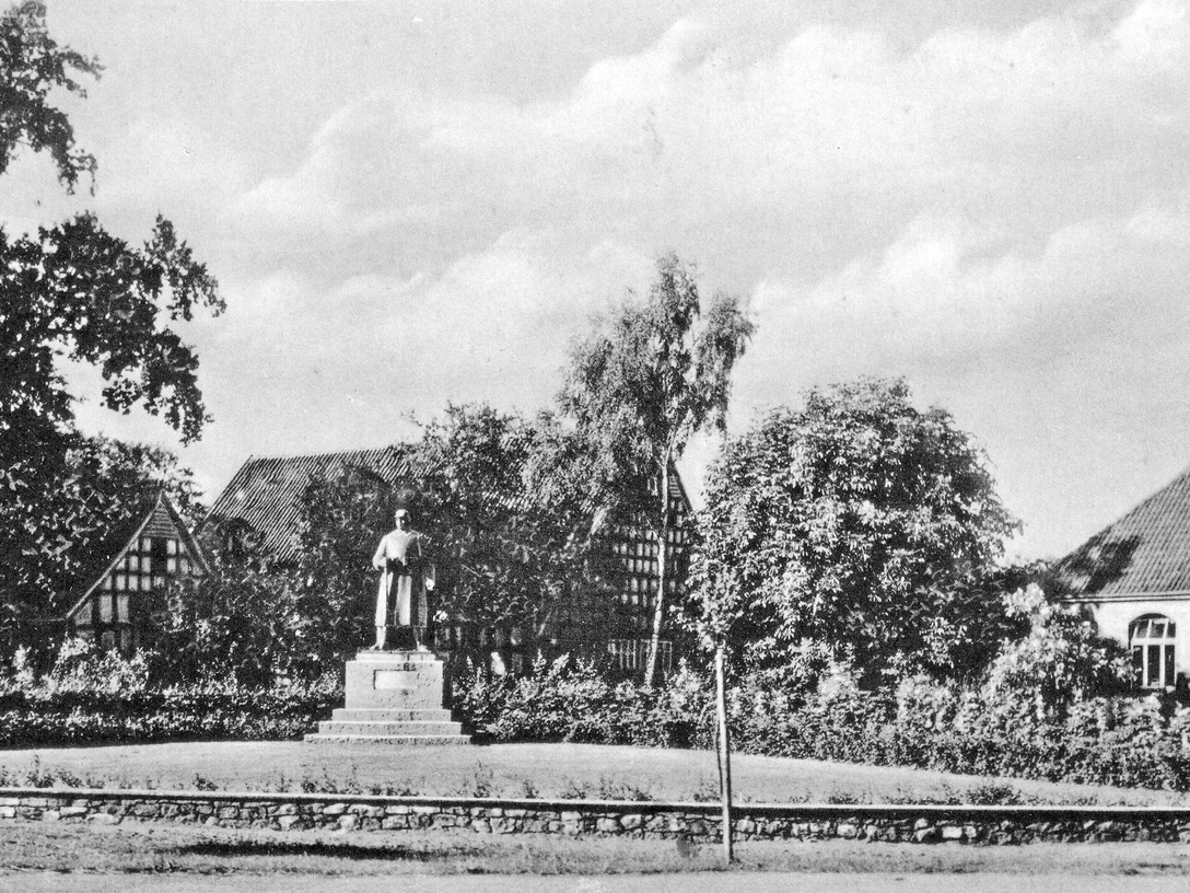 huellhorst-kriegerehrenmal-alt.jpg Kriegerdenkmal in Hüllhorst, umgeben von zwei traditionellen Fachwerkhäusern und Bäumen bei klarem Himmel.