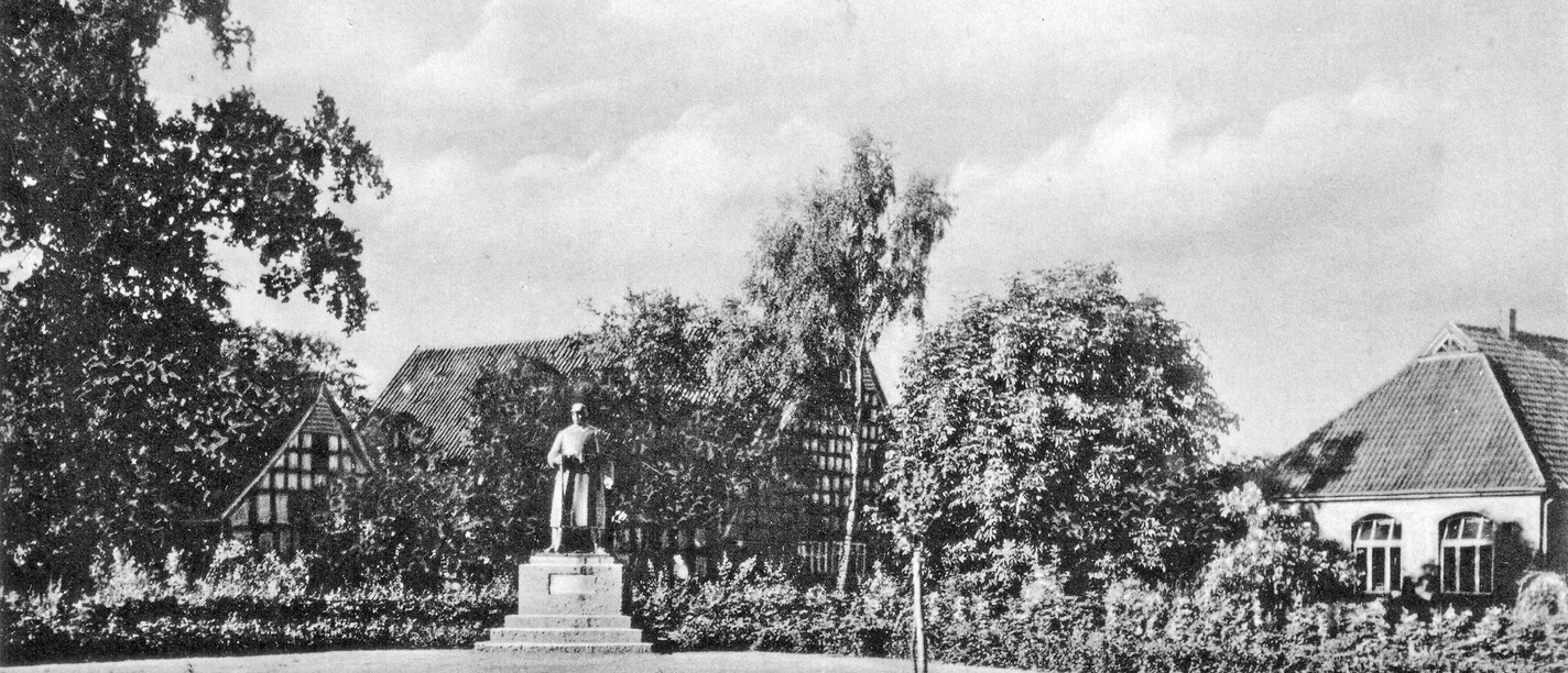 huellhorst-kriegerehrenmal-alt.jpg Kriegerdenkmal in Hüllhorst, umgeben von zwei traditionellen Fachwerkhäusern und Bäumen bei klarem Himmel.
