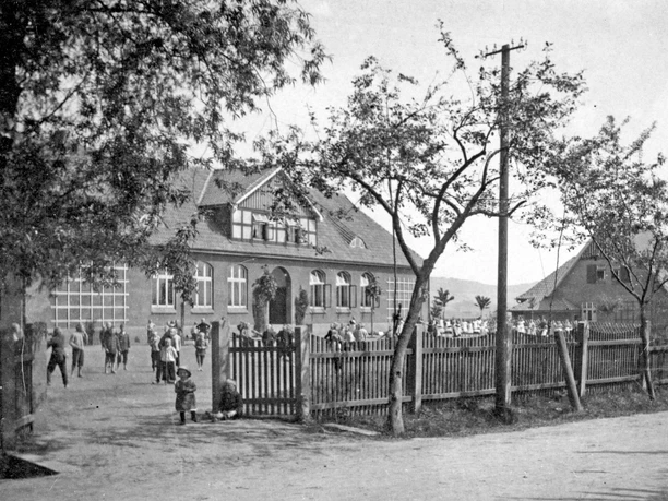 huellhorst-alte-schule-01.jpg Historisches Schulgebäude mit Ziegelsteinfassade, umgeben von Bäumen und Menschen im Vordergrund.